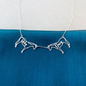 May include: A silver necklace with two horse silhouettes linked by a chain. Each horse has a name inscribed on it, "Libby" and "Joey". The necklace is shown against a dark teal fabric background.