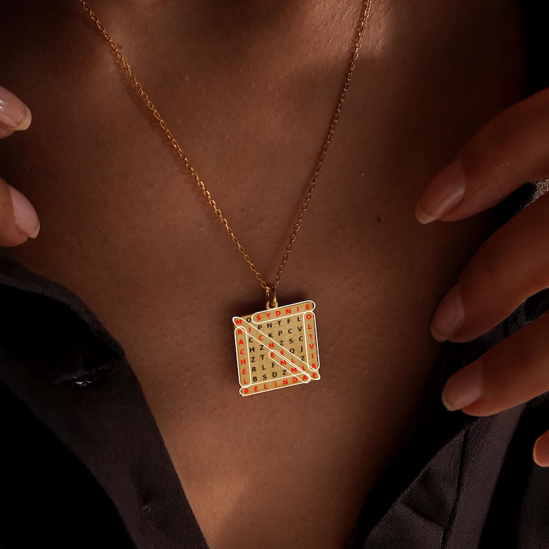 May include: Gold-toned necklace featuring a square pendant with a word search design. The pendant has a grid of letters with red accents. The necklace is displayed on a dark background.