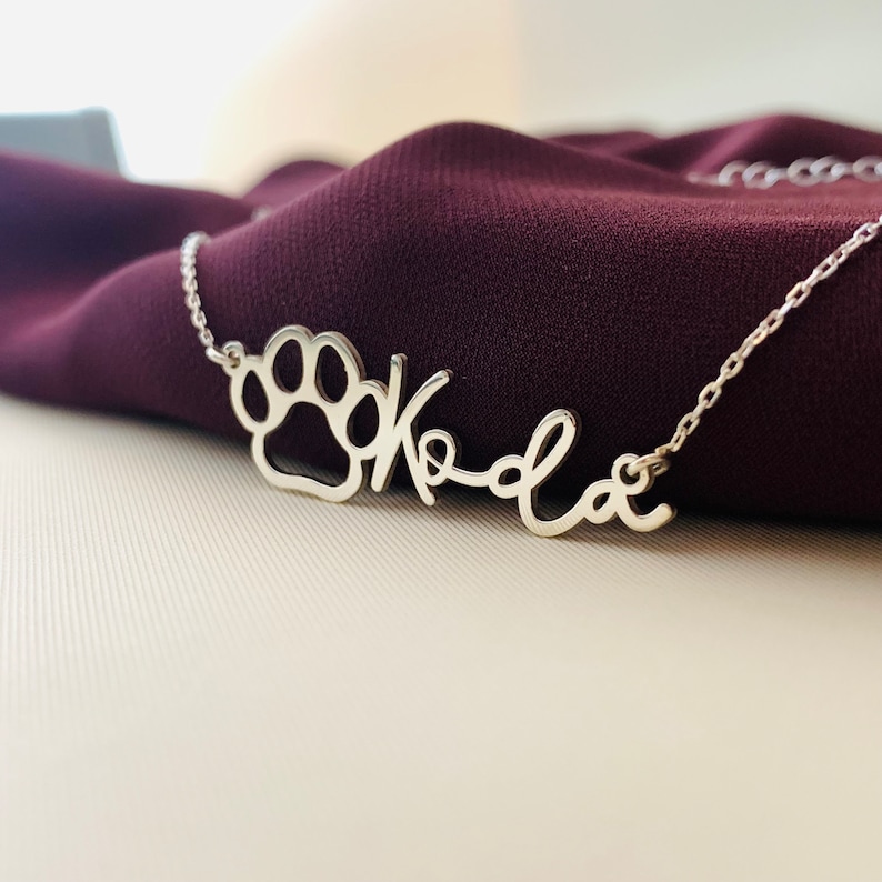 Peut inclure: Collier en argent avec un pendentif en forme de patte et le nom "Koda" &eacute;crit en cursive.