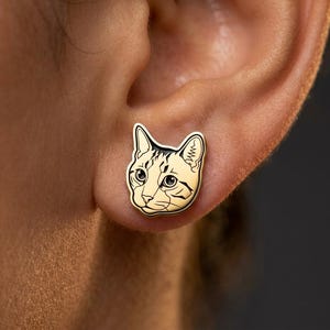 May include: A gold-toned stud earring shaped like a cat head. The earring features black enamel detailing that outlines the cat's ears, eyes, nose, and whiskers, with a smooth, polished metallic surface that captures the feline facial features.