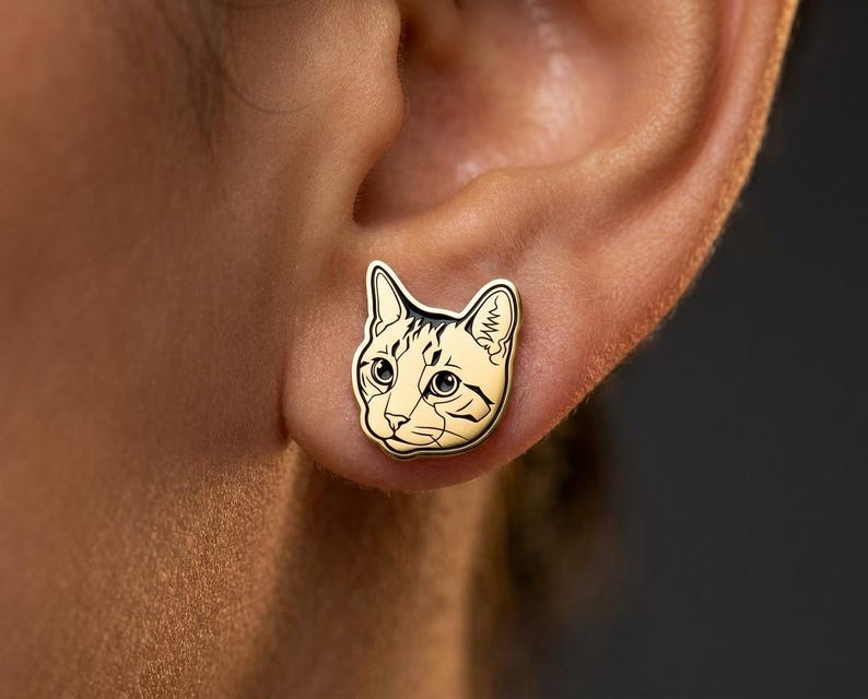 May include: A gold-toned stud earring shaped like a cat head. The earring features black enamel detailing that outlines the cat's ears, eyes, nose, and whiskers, with a smooth, polished metallic surface that captures the feline facial features.