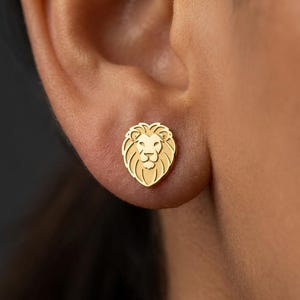 Op de afbeelding: Een gouden leeuwenoorbel. De oorbel is een gedetailleerd, klein en symmetrisch ontwerp van een leeuwenkop. De oorbel wordt op het oor gedragen. De achtergrond is een wazig beeld van de huid van de persoon.