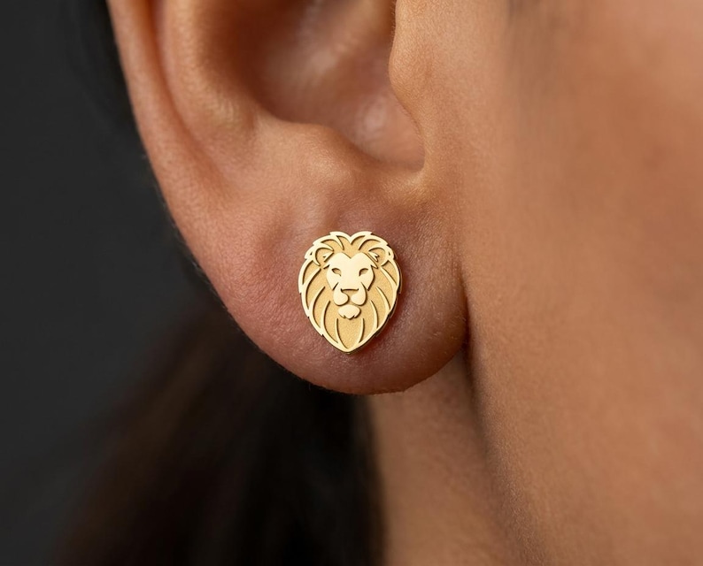 Op de afbeelding: Een gouden leeuwenoorbel. De oorbel is een gedetailleerd, klein en symmetrisch ontwerp van een leeuwenkop. De oorbel wordt op het oor gedragen. De achtergrond is een wazig beeld van de huid van de persoon.