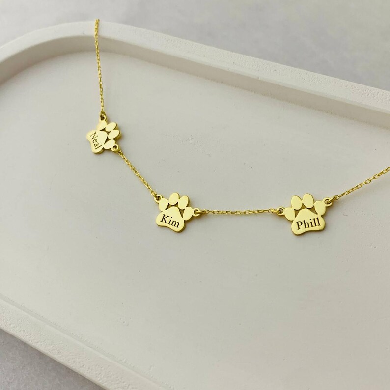 May include: Gold necklace with three paw print charms, each engraved with a name: Neil, Kim, and Phil.
