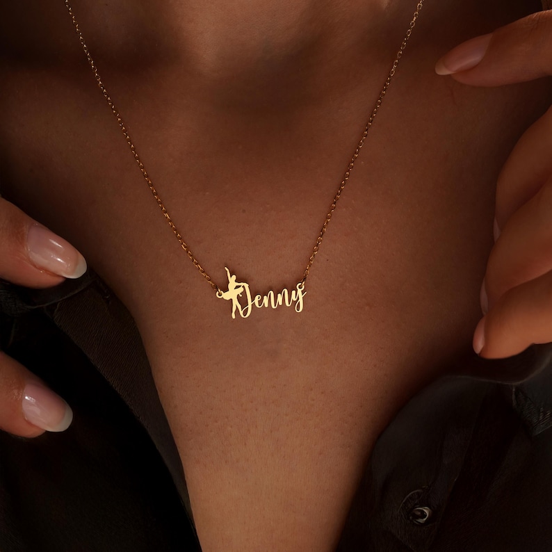 K&ouml;nnte beinhalten: Eine goldfarbene Halskette mit dem Namen "Denny" in Schreibschrift und einer Ballerina-Silhouette. Der Schmuck wird vor einem dunklen Hintergrund pr&auml;sentiert, wodurch das Design hervorgehoben wird.