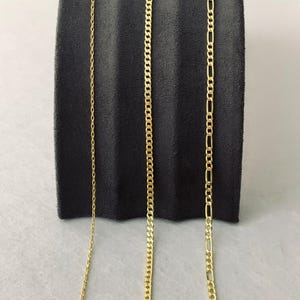 Peut inclure: Trois colliers en or pr&eacute;sent&eacute;s sur un fond gris fonc&eacute;. Les colliers pr&eacute;sentent diff&eacute;rents styles de cha&icirc;ne : une cha&icirc;ne fine, une cha&icirc;ne gourmette et une cha&icirc;ne Figaro. Les cha&icirc;nes sont pr&eacute;sent&eacute;es verticalement, mettant en valeur leurs designs individuels et leur brillance.