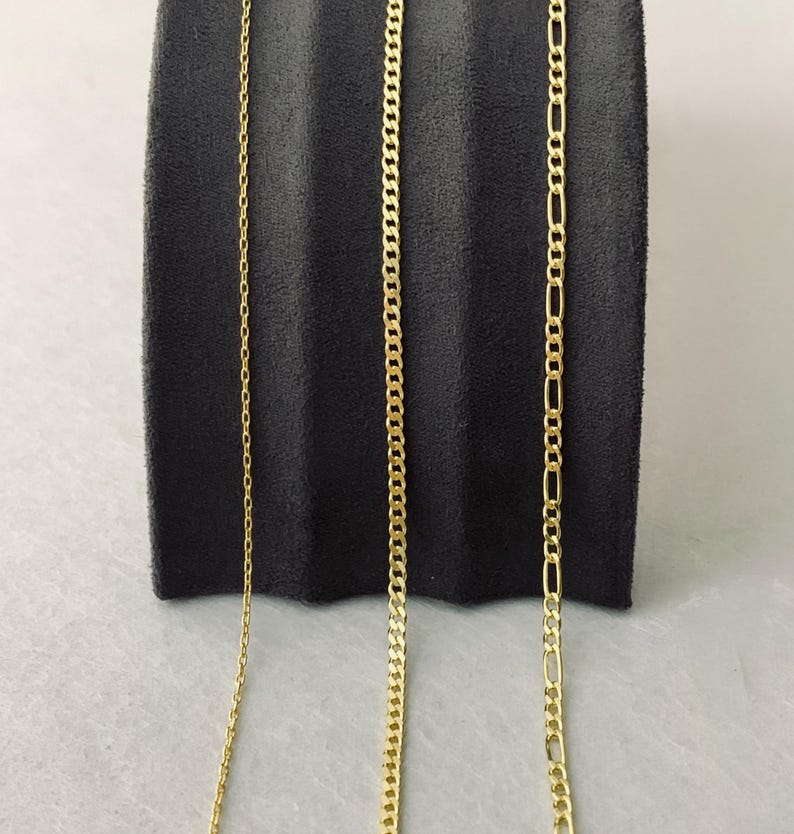 Peut inclure: Trois colliers en or pr&eacute;sent&eacute;s sur un fond gris fonc&eacute;. Les colliers pr&eacute;sentent diff&eacute;rents styles de cha&icirc;ne : une cha&icirc;ne fine, une cha&icirc;ne gourmette et une cha&icirc;ne Figaro. Les cha&icirc;nes sont pr&eacute;sent&eacute;es verticalement, mettant en valeur leurs designs individuels et leur brillance.