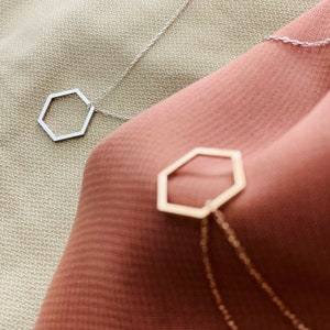 May include: Two delicate necklaces with hexagon pendant charms. One necklace is silver and the other is gold. The necklaces are displayed on a beige and brown fabric background.
