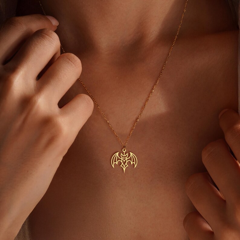 K&ouml;nnte beinhalten: Eine goldfarbene Halskette mit einem Fledermaus-Anh&auml;nger. Der Anh&auml;nger hat ein detailliertes, offenes Design mit ausgebreiteten Fl&uuml;geln und einem zentralen Emblem. Die Halskette wird um den Hals getragen, der Anh&auml;nger liegt auf der Brust.