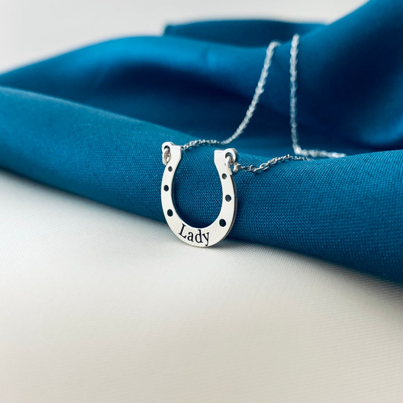 以下が含まれることがあります： 馬蹄の形をしたシルバーネックレスで、馬蹄に「Lady」という文字が刻まれています。ネックレスは青い布の背景の上に置かれています。