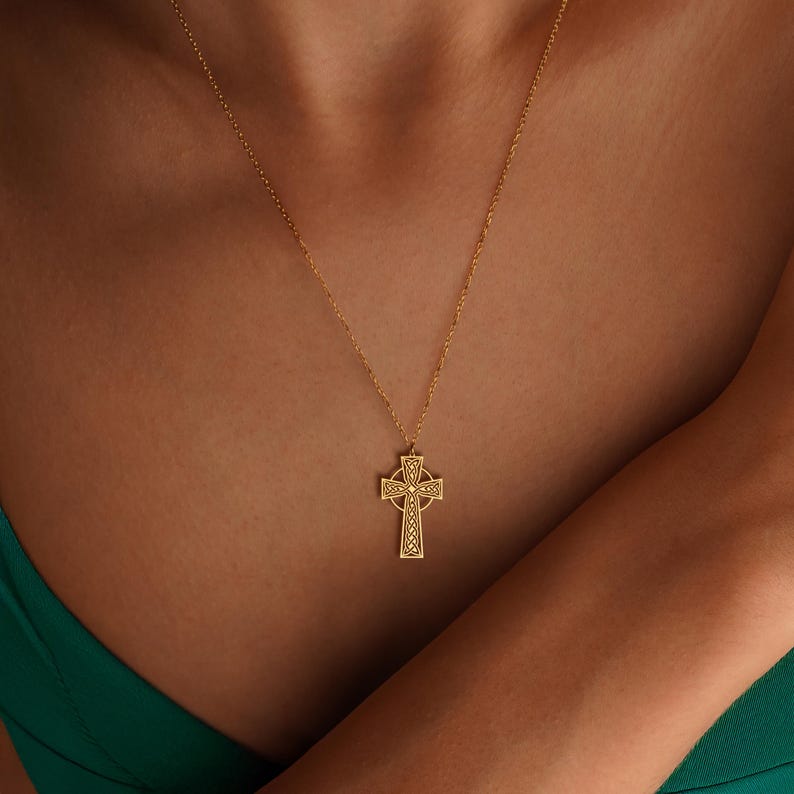 May include: A gold-coloured Celtic cross pendant necklace. The cross features intricate knotwork and is suspended from a delicate chain. The necklace is worn against a green top.