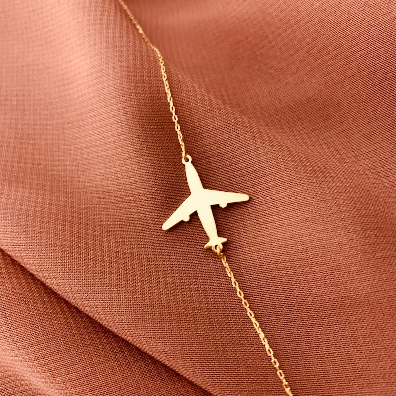 Op de afbeelding: Een gouden ketting met een delicaat vliegtuighanger. De ketting ligt op een bruine stoffen achtergrond.