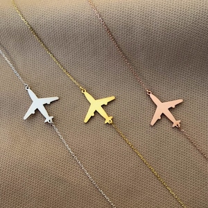 Op de afbeelding: Drie zilveren kettingen met vliegtuig hangers. De kettingen liggen op een bruine stoffen achtergrond. De vliegtuigen zijn in zilver, goud en ros&eacute; goud.