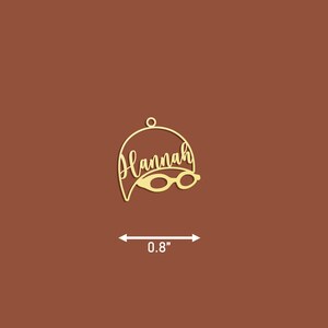 May include: Gold-coloured metal ornament shaped like a swimmer's cap with the name "Hannah" and a pair of goggles. The ornament is 0.8 inches wide.