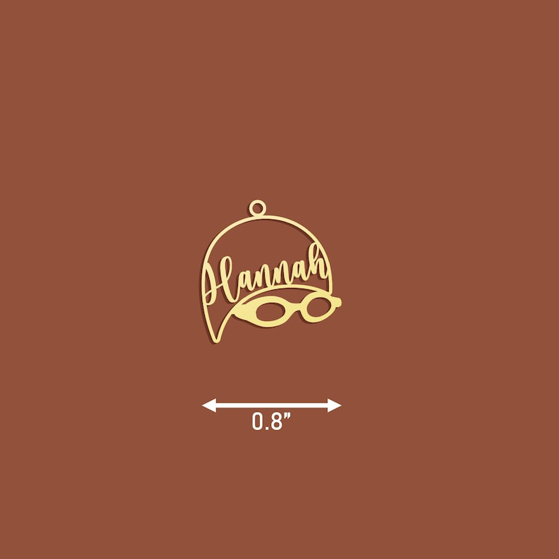May include: Gold-coloured metal ornament shaped like a swimmer's cap with the name "Hannah" and a pair of goggles. The ornament is 0.8 inches wide.