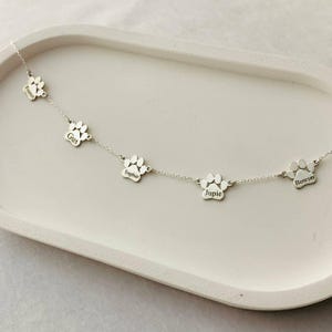 May include: Silver necklace with five paw print charms, each engraved with a different name: Frankie, Gus, Sophie, Jupie, and Bowser.