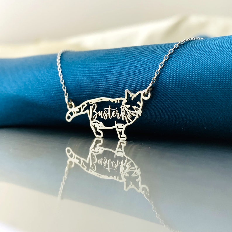 May include: A silver necklace with a cutout of a cat with the name "Buster" inscribed on it. The necklace is on a blue background.