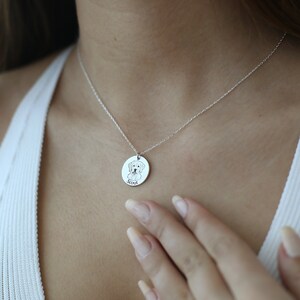 May include: A silver necklace with a circular pendant. The pendant has a dog illustration and the name "Scout" engraved below. The pendant hangs from a delicate chain. The person is wearing a white top.