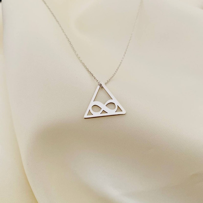 Pode incluir: Colar prateado com um pendente triangular com um s&iacute;mbolo de infinito vazado. O pendente pende de uma corrente delicada, criando um design minimalista e elegante. O colar &eacute; exibido sobre um fundo macio e esbranqui&ccedil;ado.
