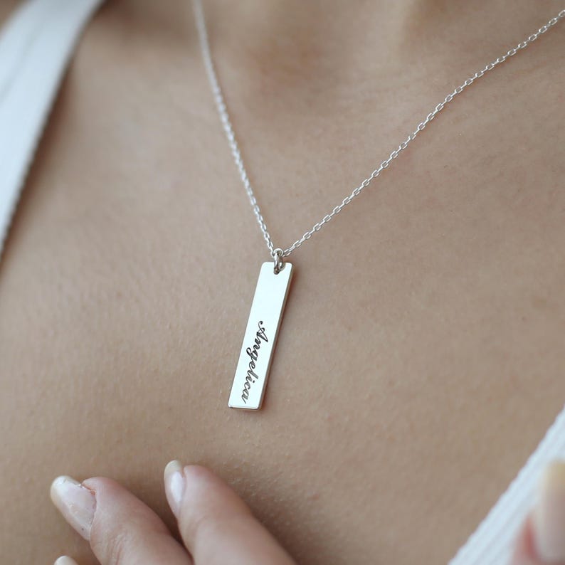 Puede incluir: Un collar de plata con un colgante rectangular que dice "Angelica" en cursiva.