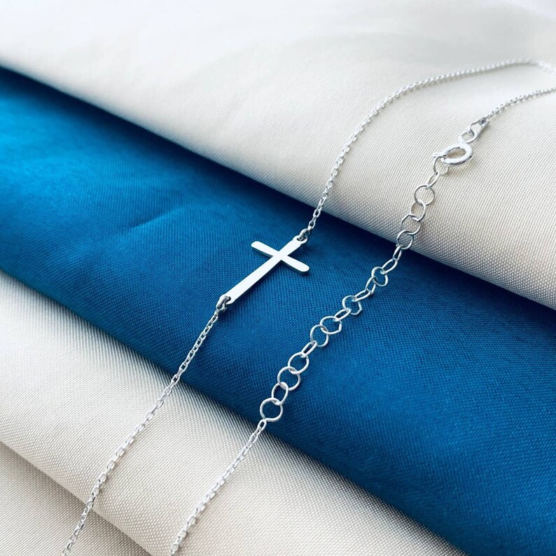 Op de afbeelding: Een zilveren ketting met een delicate zilveren kruis hanger. De ketting wordt getoond op een witte en blauwe stoffen achtergrond.
