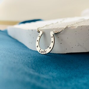 以下が含まれることがあります： 馬蹄の形をしたシルバーネックレス。馬蹄に「Lady」という文字が刻まれています。