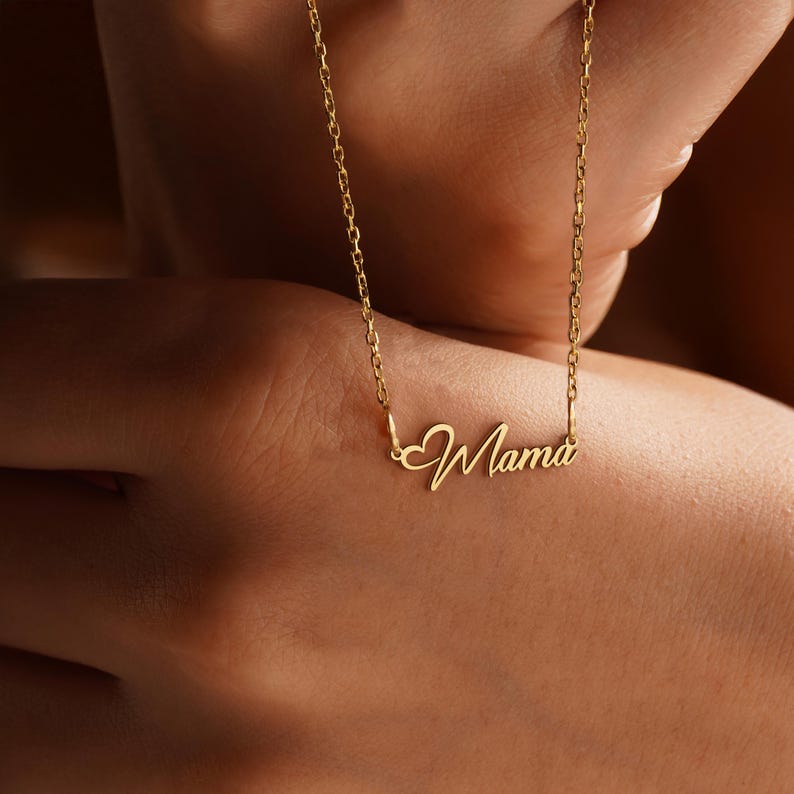 May include: A gold-colored necklace with the word "Mama" in a cursive font. The necklace has a delicate chain and a heart-shaped design element before the word. The necklace is displayed against a blurred background.