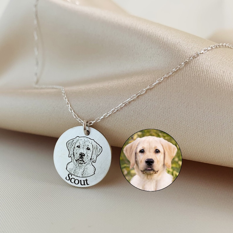 May include: A silver necklace with a circular pendant engraved with a dog portrait and the name "Scout". A small, circular photo of the dog is next to the pendant. The necklace is shown on a beige satin background.