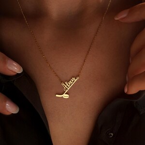 May include: A gold-colored necklace with the name "Alexis" in cursive script. The pendant design includes a hockey stick and puck. The necklace is displayed on a person wearing a dark shirt.