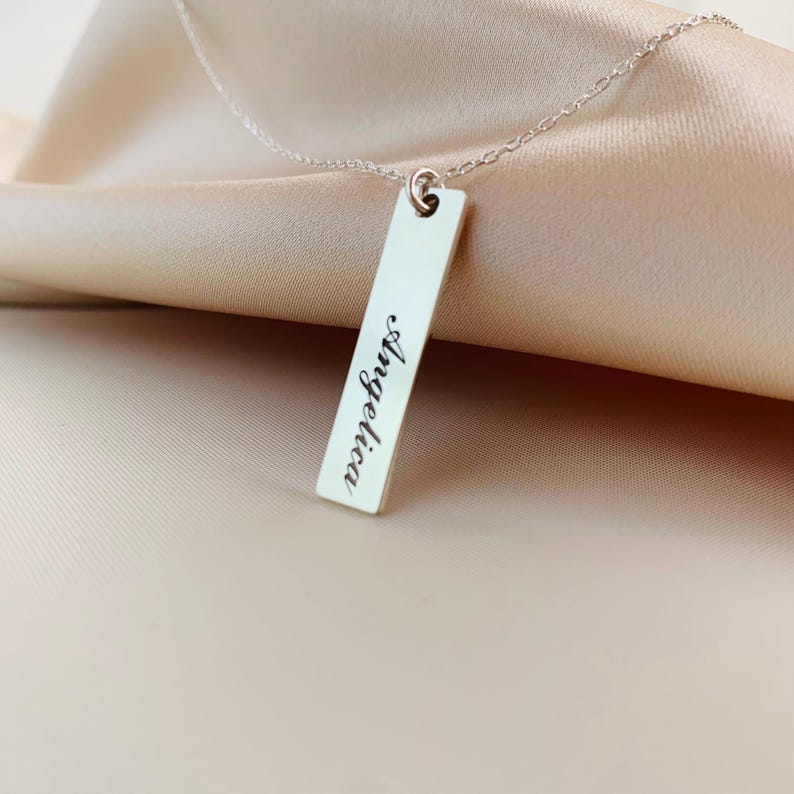Puede incluir: Un collar de plata con un colgante rectangular que dice "Angelica" en cursiva.