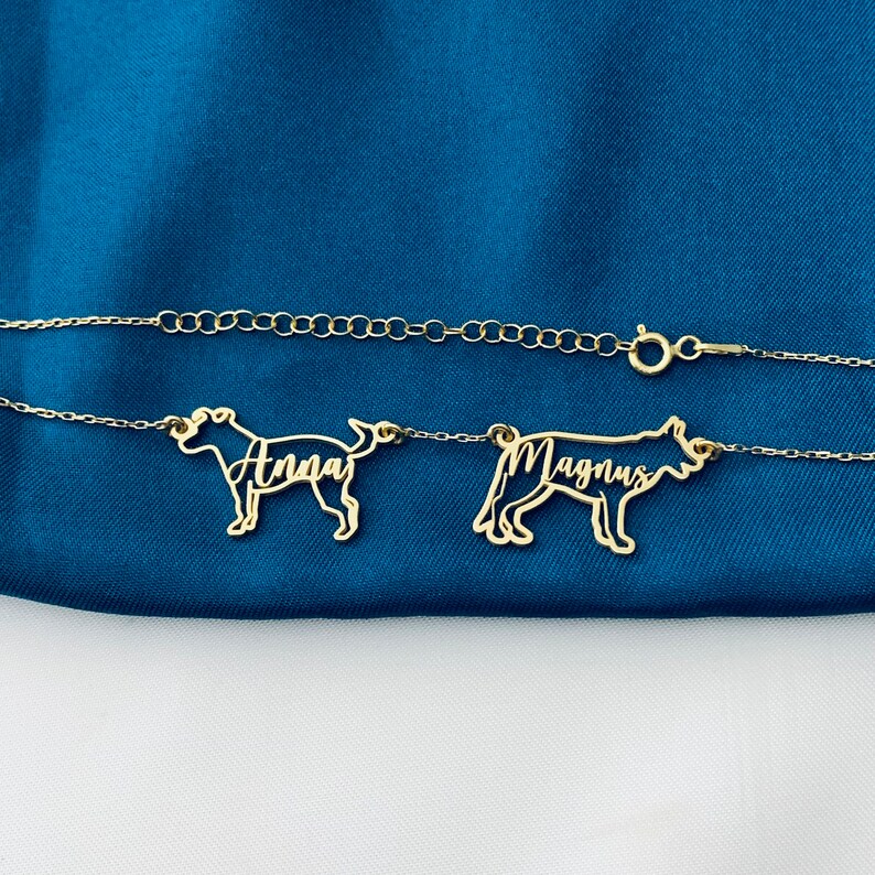 May include: A gold chain necklace with two dog-shaped pendants. The pendants are cut out and the names "Anna" and "Magnus" are written inside the cutouts.