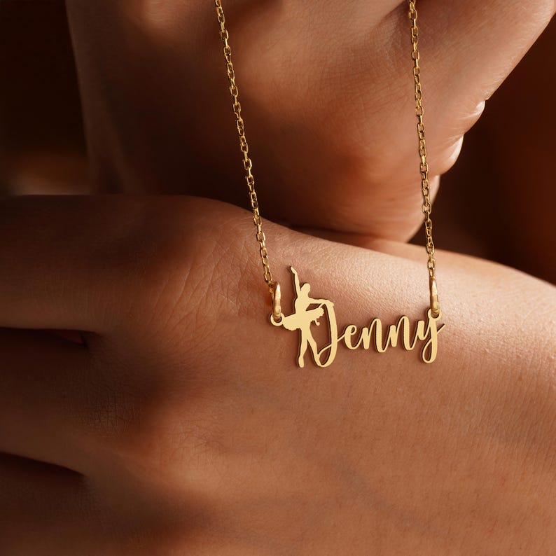 K&ouml;nnte beinhalten: Eine goldfarbene Halskette mit feiner Kette. Der Anh&auml;nger zeigt den Namen "Jenny" in Schreibschrift, mit einer Ballerina-Silhouette in das Design integriert. Die Halskette wird vor einem unscharfen Hintergrund gezeigt.