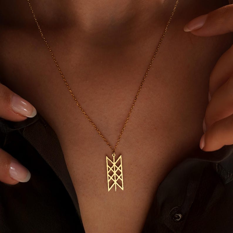 May include: A gold necklace featuring a geometric pendant. The pendant is rectangular with an open, symmetrical design. The necklace is shown against a dark backdrop.