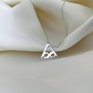 Pode incluir: Um colar de prata com um pingente triangular com um s&iacute;mbolo de infinito vazado. O pingente pende de uma corrente delicada. O colar &eacute; exibido em um tecido branco e sedoso, criando uma est&eacute;tica minimalista.