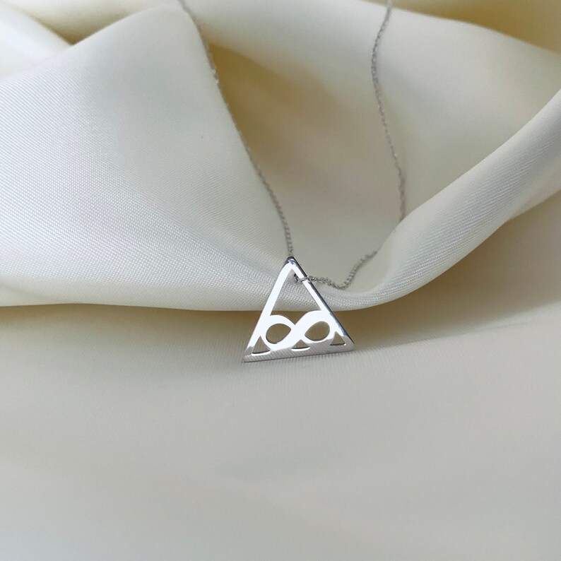 Pode incluir: Um colar de prata com um pingente triangular com um s&iacute;mbolo de infinito vazado. O pingente pende de uma corrente delicada. O colar &eacute; exibido em um tecido branco e sedoso, criando uma est&eacute;tica minimalista.
