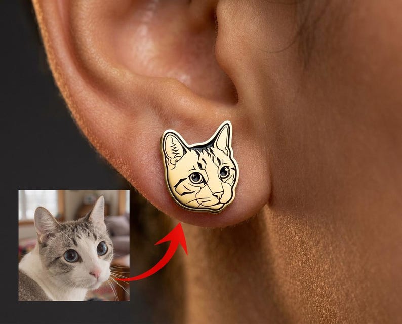 May include: A gold-toned stud earring shaped like a cat head with black engraved line details. The earring features a stylised illustration of a cat face with pointed ears, whiskers, and large eyes, matching the cat shown in the small inset photo.