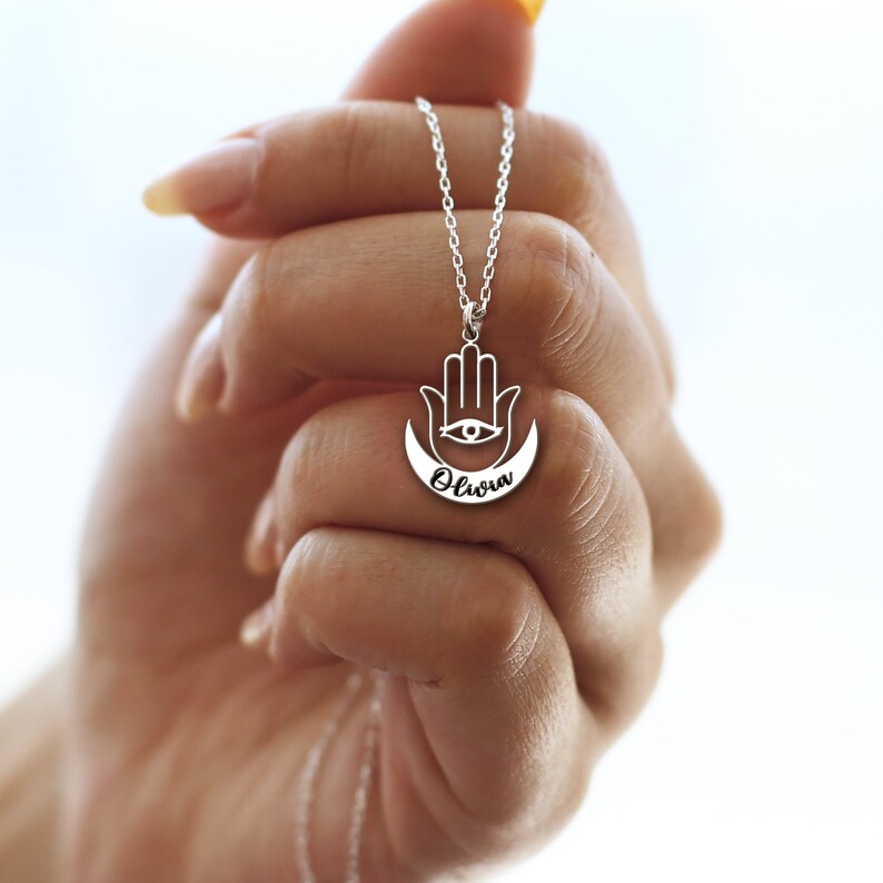 May include: Silver necklace with a hamsa hand charm and a crescent moon with the name "Olivia" inscribed on it.