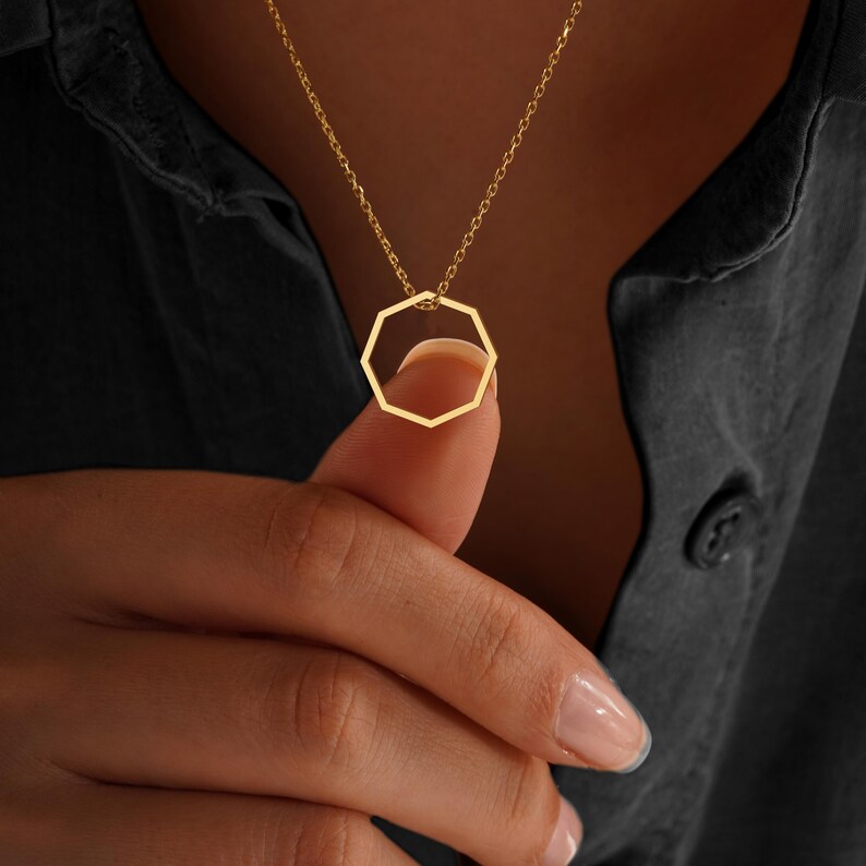 Peut inclure: Un collier dor&eacute; avec un pendentif g&eacute;om&eacute;trique. Le pendentif est une forme ouverte &agrave; plusieurs c&ocirc;t&eacute;s, suspendue &agrave; une fine cha&icirc;ne. Le collier est pr&eacute;sent&eacute; sur un fond sombre, mettant en valeur le design du bijou.