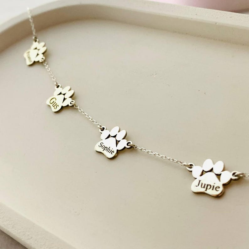 May include: Silver necklace with four paw print charms, each engraved with a different name: Gus, Sophie, and Jupie.