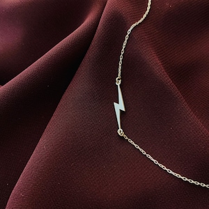 Puede incluir: Un collar de plata con un colgante de rayo en un fondo de tela color burdeos.