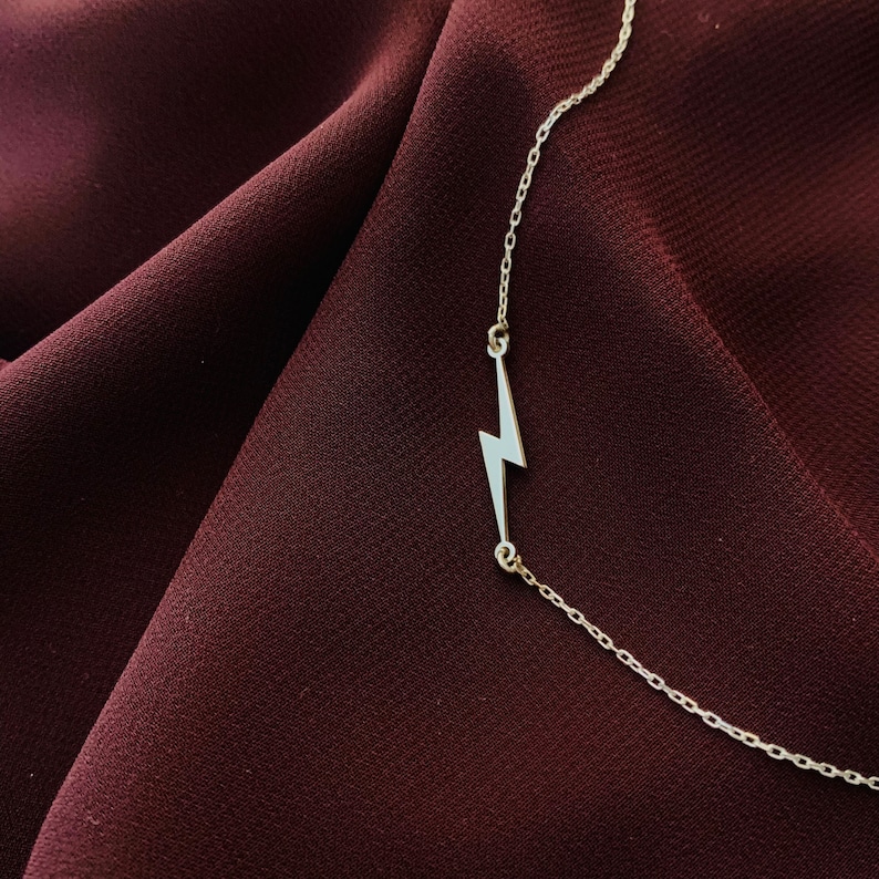 Puede incluir: Un collar de plata con un colgante de rayo en un fondo de tela color burdeos.