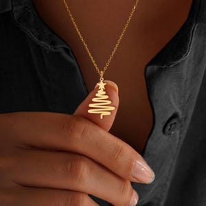 May include: A gold-colored necklace with a pendant shaped like a Christmas tree. The tree design is a stylized, continuous line with a star at the top. The necklace is on a gold chain, and the pendant is held by a person.