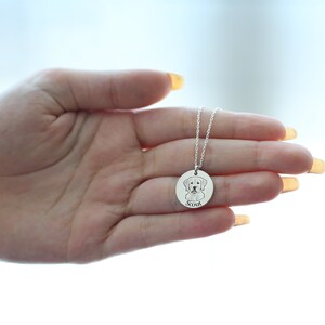 May include: A silver pendant necklace with a detailed engraved image of a dog's face. The dog's name, "Scout," is engraved below the image. The necklace is held in a hand against a bright background.