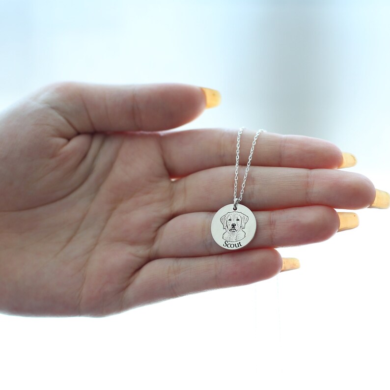 May include: A silver pendant necklace with a detailed engraved image of a dog's face. The dog's name, "Scout," is engraved below the image. The necklace is held in a hand against a bright background.