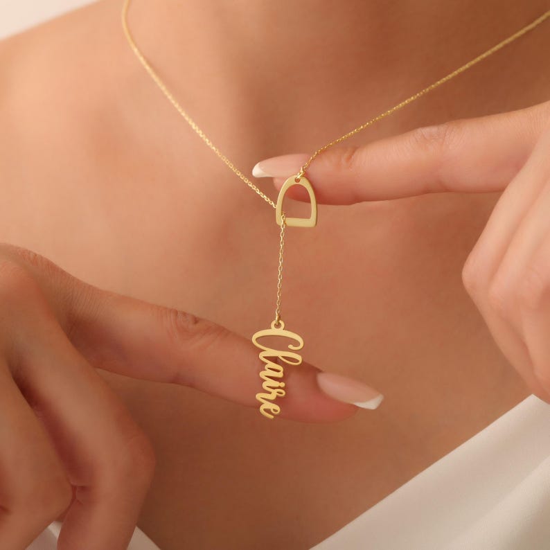 May include: A gold-colored necklace with a unique design. The necklace features a stirrup-shaped pendant connected to a chain, with the name "Claire" written in cursive script hanging below. The necklace is displayed against a neutral background.