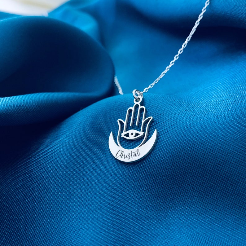 May include: Silver necklace with a hamsa hand charm and a crescent moon. The charm has an eye in the center and the name "Cristal" is engraved on the moon.