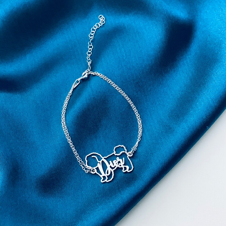 May include: A silver chain bracelet featuring a cut-out design of a dog with the name "Duer" inscribed on it. The bracelet is displayed against a blue fabric backdrop.