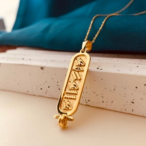 May include: A gold necklace with a pendant featuring an Egyptian hieroglyphic cartouche. The cartouche is a rectangular shape with a winged scarab beetle, an eye of Horus, and other hieroglyphs.