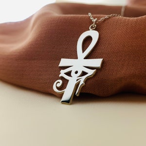 Peut inclure: Un collier pendentif argent&eacute; avec un design Ankh et &OElig;il d'Horus. Le pendentif est suspendu &agrave; une fine cha&icirc;ne, sur un fond de tissu marron. Le collier est un symbole de la culture &eacute;gyptienne ancienne.