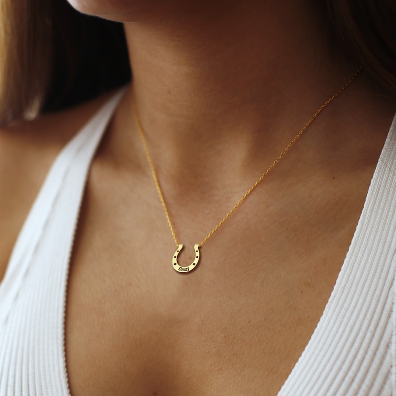 K&ouml;nnte beinhalten: Eine goldene Hufeisenkette mit dem Wort "Daisy" auf dem Hufeisen. Die Kette tr&auml;gt ein Model mit einem wei&szlig;en Tanktop.